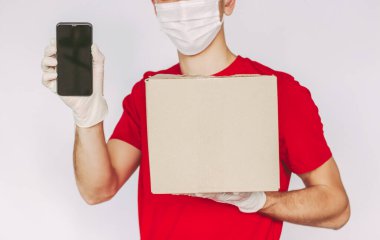 Tıbbi yüz maskesi takan, yakın plan teslimatçı, koruyucu eldiven akıllı telefon, elinde karton kutu, boş fotokopi alanı. Teslimatçı, paket kutusu, cep telefonu, izole bir geçmiş taşıyor.