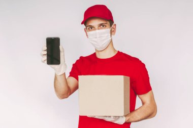 Portre mutlu servis elemanı tıbbi yüz maskesi, koruyucu eldivenler cep telefonu, boş fotokopi alanı olan paket kutusu. Genç teslimatçı karton kutu ve akıllı telefon taşıyor.