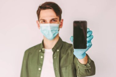 Koruyucu eldiven ve tıbbi maske takan genç adamın portresi beyaz arka planda boş fotokopi alanı olan akıllı telefonu tutuyor. Elinde cep telefonu olan mutlu bir adam. Dijital mobil kişi daha az ödeme