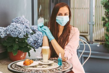 Fransız kafe 'sinde koruyucu maskeli ve tıbbi eldivenli güzel mutlu bir kız. Tıbbi maske ve koruyucu eldiven takan genç bir kadın, şehir caddesindeki restoran terasında otururken kahve içer.