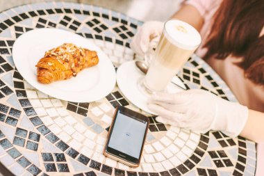 Yakın plan, koruyucu eldivenli genç bir kadın kahve içip Fransız kahvesinde kruvasan içiyor. Açık hava restoranının terasında tıp eldivenli güzel bir kız. Ekranda NFC cep telefonu ödemesi olan akıllı telefon