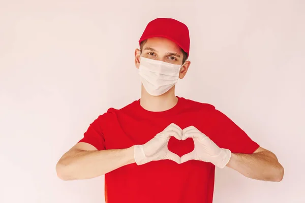 Tıbbi maske takan portre teslimatçısı, eldivenler, beyaz arka planda izole edilmiş elleri olan aşk işareti. Koruyucu maskeli mutlu teslimatçı aşk sembolü gösteriyor. Müşteri hizmetleri