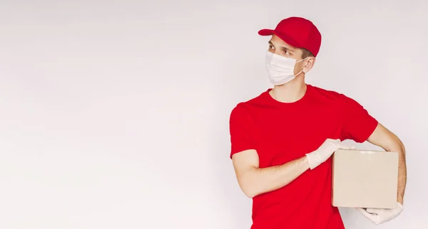 Happy delivery guy in red uniform, protective gloves, medical face mask carry cardboard box in hands, look at copy space on white background. Young man delivery service employee with shipment package