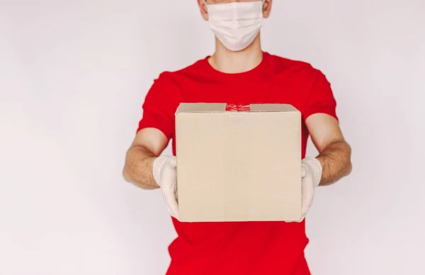 Closeup young man delivery service courier in uniform, medical face mask, protective gloves carry cardboard box in hands isolated on white background. Delivery guy give parcel shipment. Safe delivery