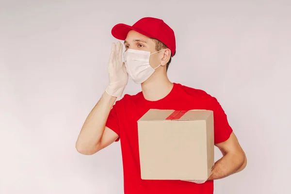 Portrait happy man delivery service employee in uniform, medical face mask, protective glove shout with hand, hold cardboard box isolated on white background. Young delivery guy make loud announcement