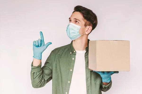 Portrait young man delivery service worker in medical face mask and protective gloves point index finger up to copy space on white background. Happy delivery guy carry cardboard parcel box in hand