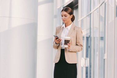 Şık takım elbiseli güzel iş kadını portresi akıllı telefon kullanır ve dışarıda kahve içer. Çekici kız profesyonel girişimci elinde kağıt kahve fincanı ve cep telefonu tutuyor. Yemek molası.