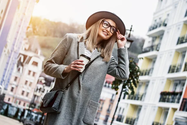Coffee on the go. Low-angle view of stylish young woman in coat and hat adjusting eyeglasses and smiling while drinking coffee outdoors. Fashionable girl walking in city. Urban fashion concept.