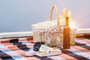 Şarap şişesi, şarap kadehi, piknik sepeti ve açık havada kareli battaniyede peynir dilimli üzümler. Romantik Fransız pikniği. Tahtada sarı ve mavi peynir. Peynirli ve üzümlü kırmızı şarap.
