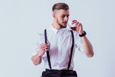 Modern saç stili olan zarif, şık bir adam ve klasik olarak gri arka planda dikilirken viski ya da viski içiyor. Tişörtlü, pantolon askılı, elinde bir bardak alkolle şık bir adam.