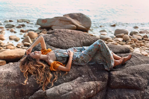 Bela jovem mulher em vestido elegante deitado em pedra ao pôr do sol — Fotografia de Stock