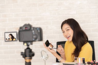 Gülümseyen Asyalı güzellik etkileyicisi, masasında çeşitli kozmetik ürünleri olan bir makyaj ürününü kameraya sunan bir video kaydeder..