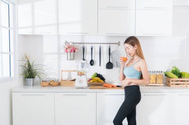 Mavi aktif giysili genç Asyalı kadın temiz ve modern mutfakta sağlıklı yaşam için bir bardak ev yapımı meyve ve sebze suyu yudumluyor..