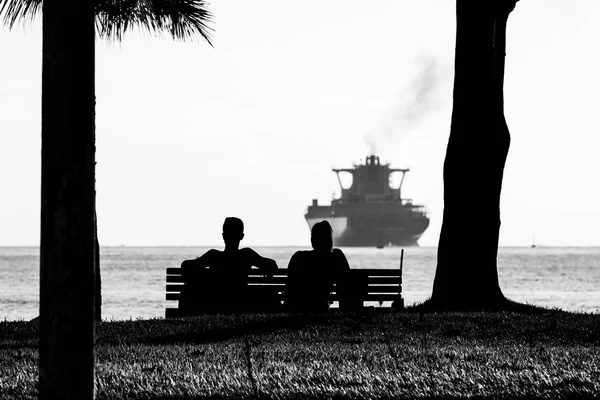 Silhouette of couple sitting on a bench at the edge of the beach watching a cargo ship pulling away.