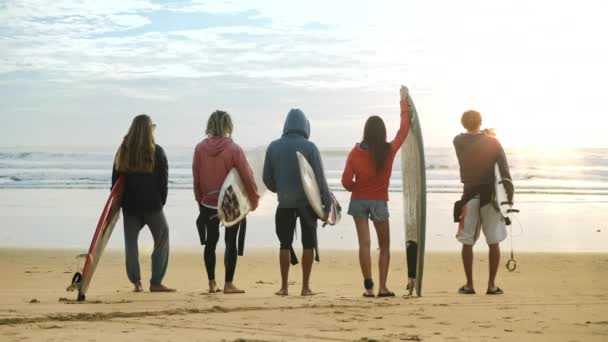 Un grupo de jóvenes surfistas sosteniendo tablas de surf, observando las olas del océano — Vídeo de stock