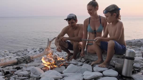 Familia feliz disfrutando de la puesta de sol de verano en la playa salvaje alrededor de la hoguera . — Vídeos de Stock