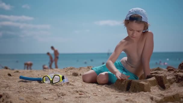 Улыбающийся мальчик играет с песком на пляже, берет маску для дайвинга, идет нырять в море — стоковое видео