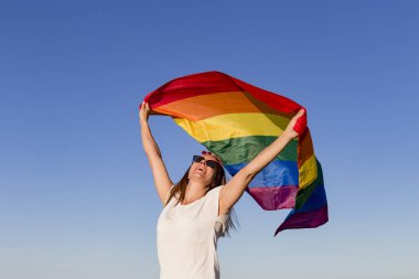 Açık havada mavi gökyüzü üzerinde Gay Gökkuşağı Bayrağı tutan kadın. Aynı cinsiyetli çiftler için mutluluk, özgürlük ve aşk konsepti. Yaşam tarzı açık havada