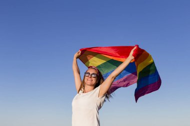 Açık havada mavi gökyüzü üzerinde Gay Gökkuşağı Bayrağı tutan kadın. Aynı cinsiyetli çiftler için mutluluk, özgürlük ve aşk konsepti. Yaşam tarzı açık havada