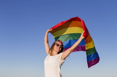 Açık havada mavi gökyüzü üzerinde Gay Gökkuşağı Bayrağı tutan kadın. Aynı cinsiyetli çiftler için mutluluk, özgürlük ve aşk konsepti. Yaşam tarzı açık havada