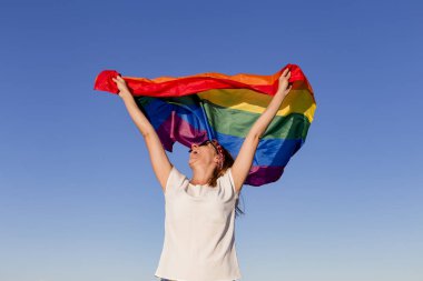 Açık havada mavi gökyüzü üzerinde Gay Gökkuşağı Bayrağı tutan kadın. Aynı cinsiyetli çiftler için mutluluk, özgürlük ve aşk konsepti. Yaşam tarzı açık havada