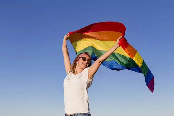 Açık havada mavi gökyüzü üzerinde Gay Gökkuşağı Bayrağı tutan kadın. Aynı cinsiyetli çiftler için mutluluk, özgürlük ve aşk konsepti. Yaşam tarzı açık havada