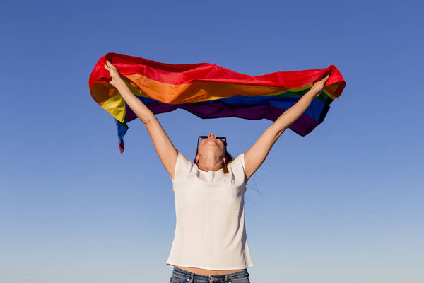Woman holding the Gay Rainbow Flag over blue sky outdoors. Happiness, freedom and love concept for same sex couples. LIfestyle outdoors