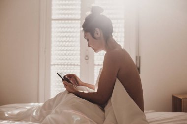 çıplak kadın yatakta oturan ve bir fincan kahve zevk beyaz çarşaf ile kaplı. Güneş pencereden geliyor. İç yaşam tarzı