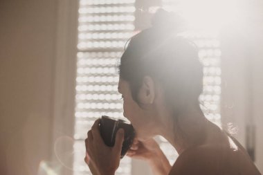 çıplak kadın yatakta oturan ve bir fincan kahve zevk beyaz çarşaf ile kaplı. Güneş pencereden geliyor. İç yaşam tarzı