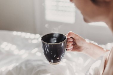çıplak kadın yatakta oturan ve bir fincan kahve zevk beyaz çarşaf ile kaplı. Güneş pencereden geliyor. İç yaşam tarzı