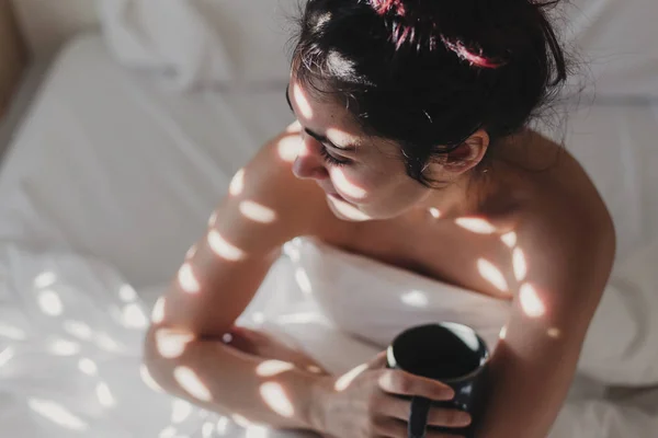 naked woman covered with white sheets sitting on bed and enjoying a cup of coffee. sun is coming through the window. Indoors lifestyle