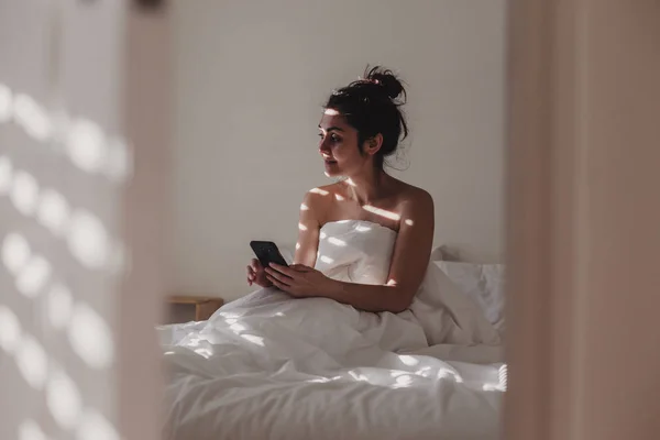 Wanita Ditutupi Dengan Seprai Putih Duduk Tempat Tidur Dengan Telepon — Stok Foto