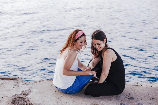 Dua Teman Wanita Yang Duduk Pantai Menggunakan Ponsel Dan Tersenyum — Stok Foto