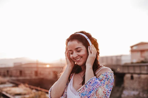 portrait outdoors of a young beautiful woman at sunset listening music on headset and smiling. Lifestyle and music concept