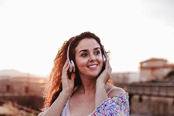 portrait outdoors of a young beautiful woman at sunset listening music on headset and smiling. Lifestyle and music concept
