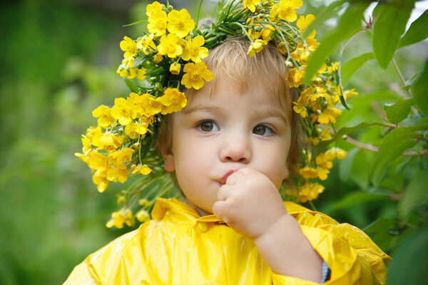 A small child in a yellow raincoat and a wreath of buttercups eats berries from a bush.