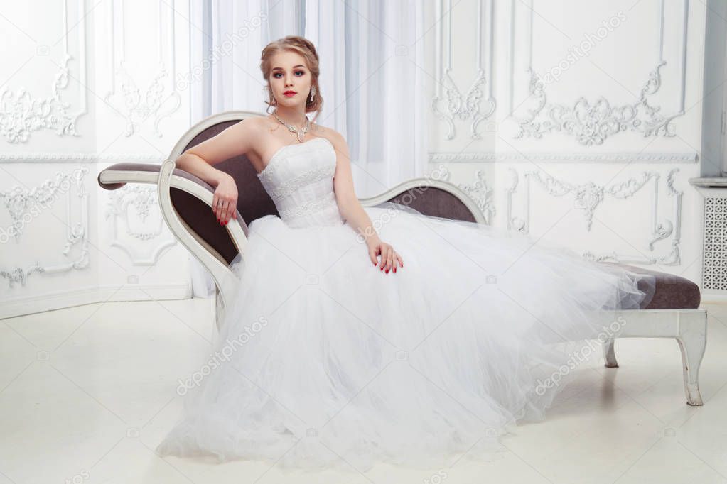 A sweet bride in a beautiful dress sitting  in an armchair in a chic, light interior.
