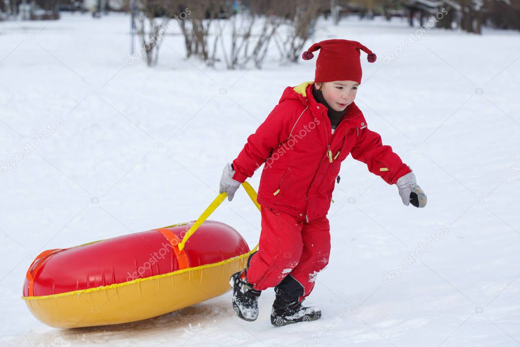 A child in a red jumpsuit rolls tubing uphill in a winter park.