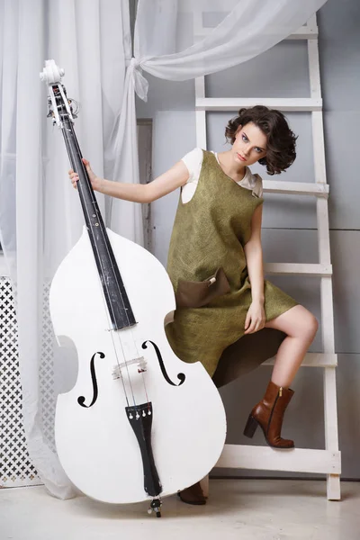 young woman in green dress with double bass standing in room with white ladder