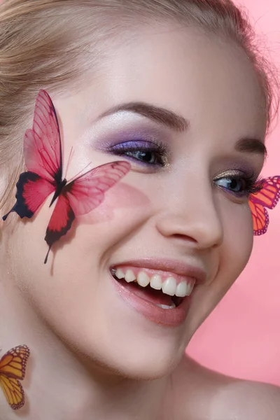 Beauty fashion portrait of a beautiful smiling girl with a gentle make-up and butterflies on her face isolated on a pink background.