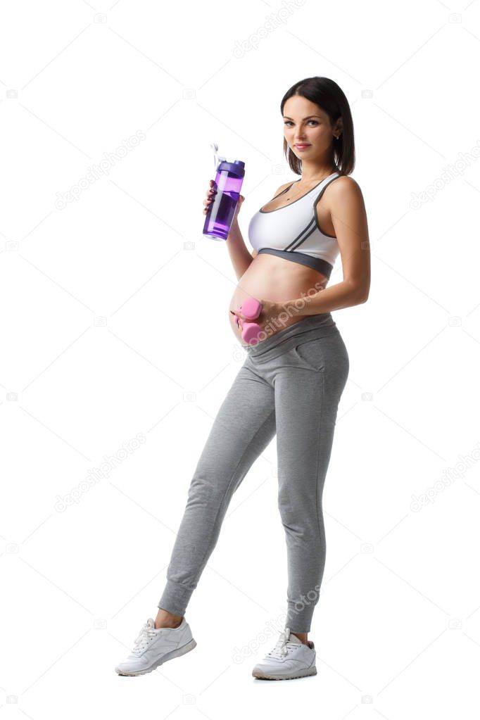Slim athletic pregnant girl is standing with a bottle of water and a dumbbell in hand isolated on white background.