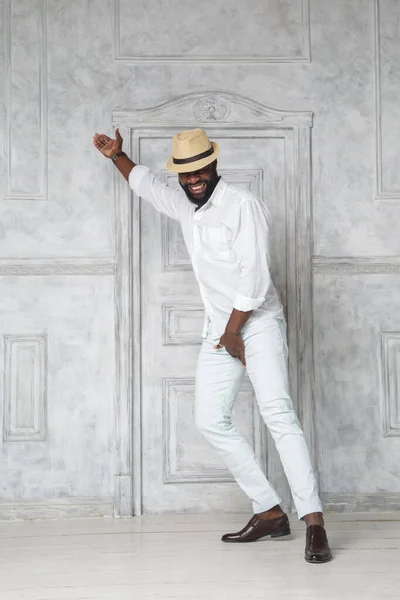 Black elegant man in a white suit and hat is dancing in a bright room near the door.