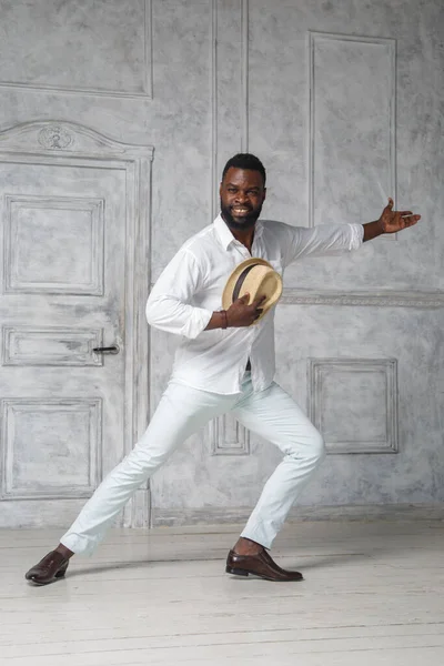 Black elegant man in a white suit is dancing in a bright room near the door holding a hat in one hand.