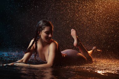 Yüzüne damla damla su damlatan güzel seksi bir kız. Stüdyonun arka planında turuncu bir ışıkla aydınlatılmış..
