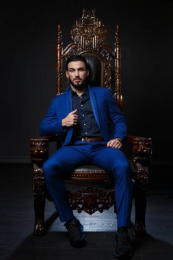 Klasik mavi takım elbiseli şık bir adam oyulmuş ahşap bir sandalyede koyu bir arka planda oturuyor..