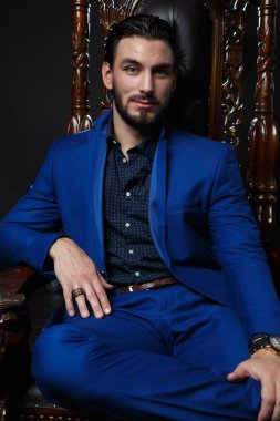 Klasik mavi takım elbiseli şık bir adam oyulmuş ahşap bir sandalyede koyu bir arka planda oturuyor..