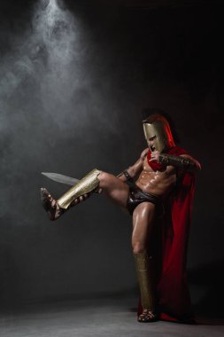Kırmızı pelerinli Romalı bir savaşçı gibi giyinmiş genç bir atletik ve siyah arka planda bir kask, esrarengiz dumanlarla örtülü, bir kılıçla dövüş tekniklerini gösteriyor..