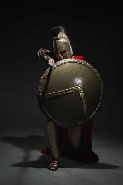 Kırmızı pelerinli Romalı asker gibi giyinmiş genç bir atletik ve siyah arka planda bir kask bir dizinin üzerine çöktü ve bir mızrağı hedef alarak kendisini kalkan ile kapladı..