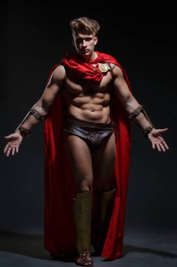 Siyah arka planda kırmızı pelerinli Roma savaşçısı kostümlü genç atletik bir adam elini bir karşılama jesti ile uzattı..