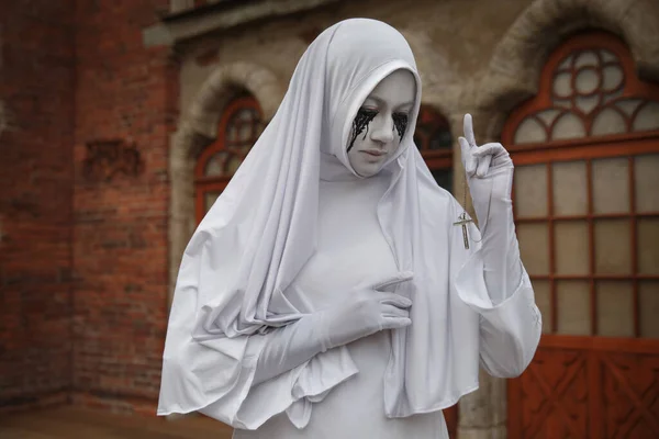 A gloomy portrait of a ghostly pale nun in white robes with black eye sockets and black smudges under them, amid gloomy Gothic architecture. Halloween, horror.